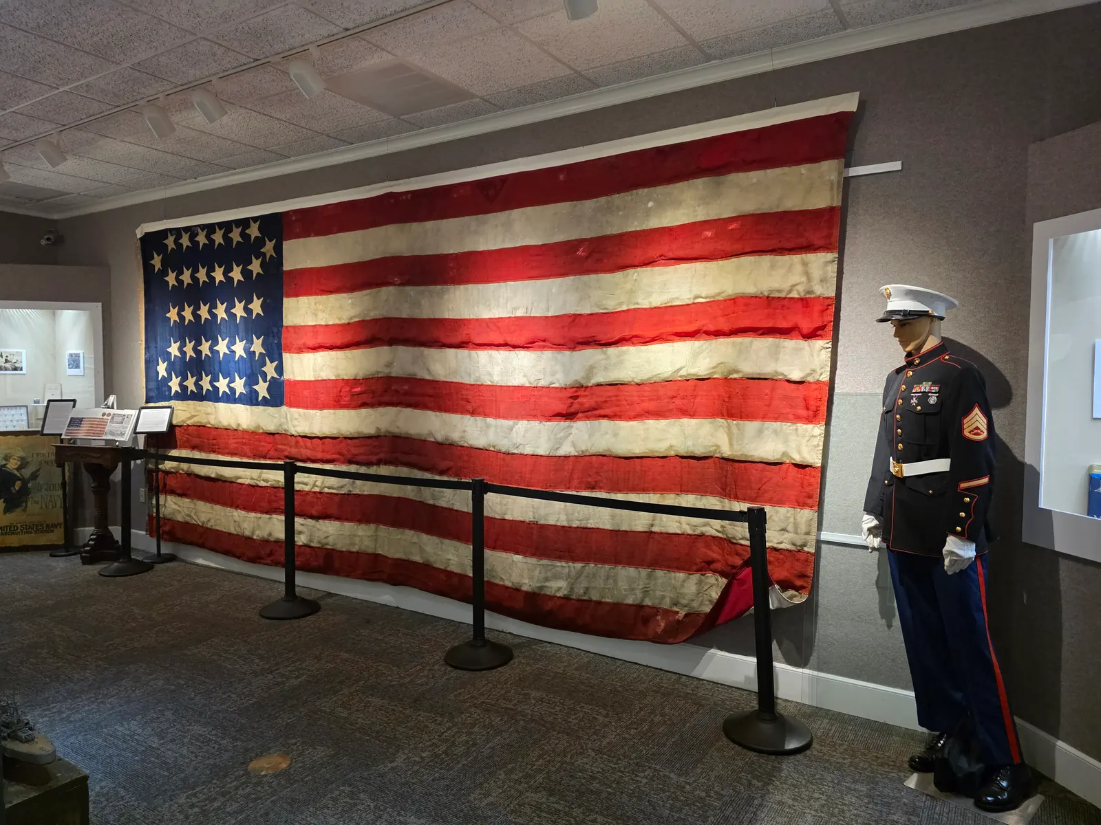 A large, worn Civil War–era American flag spans a gallery wall, beside a Marine mannequin in dress blues