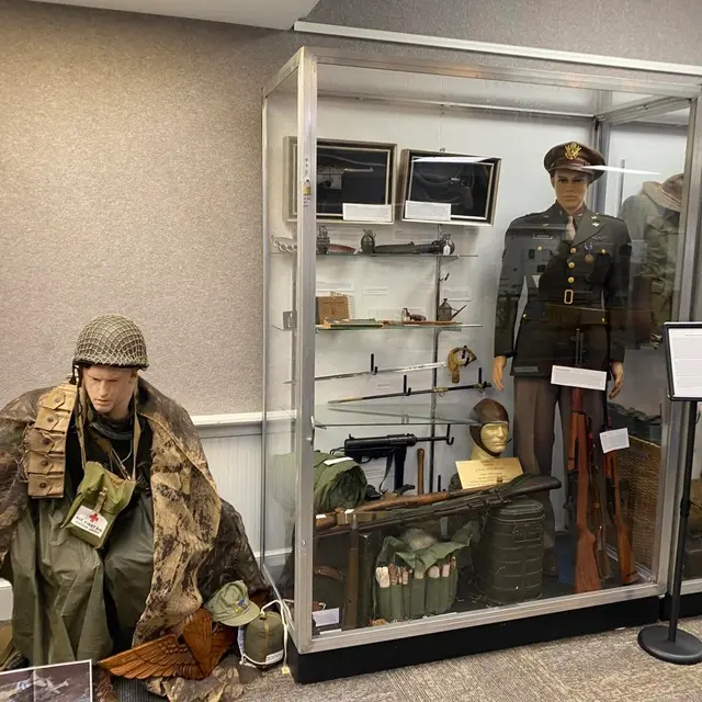 A mannequin in a helmet and camouflage cape sits beside a glass case holding an officer’s uniform, a submachine gun, rifles and field gear