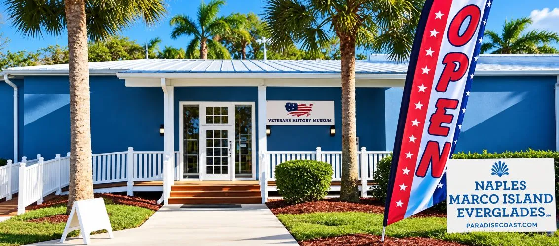 The Veterans History Museum entrance: a blue building with a white porch and railings among palm trees, with an OPEN flag and a Paradise Coast sign out front